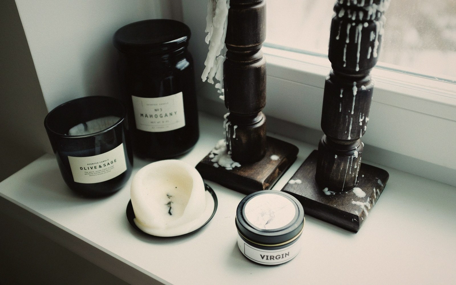 Labeled candle jars and a wax-covered tin arranged beside melted wax-drenched wooden candlesticks