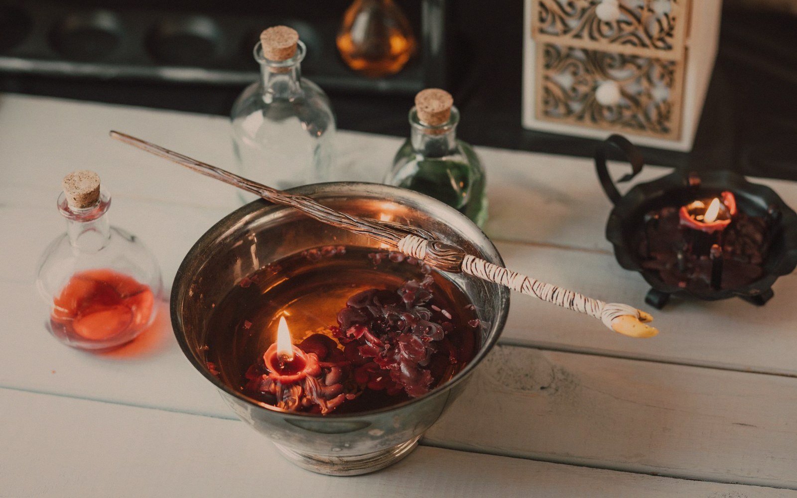 A wand rests across a metal bowl holding a lit candle and dried flowers, surrounded by corked glass bottles