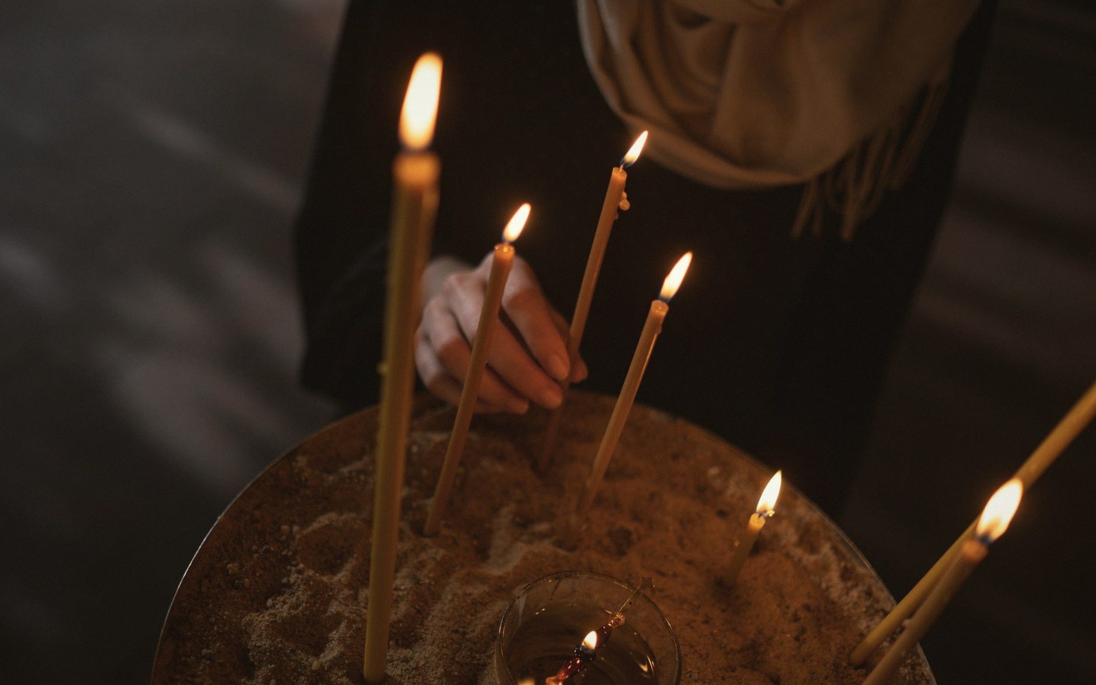 A person places a lit taper candle into a sand-filled tray among others