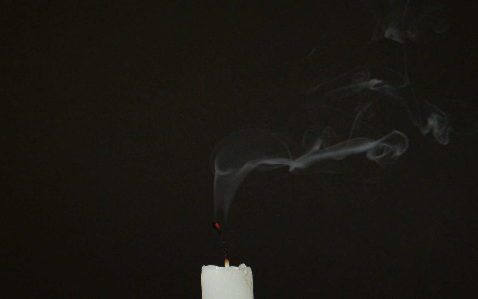 A just-extinguished candle wick trailing a thin line of smoke against a dark warm background, the wax surface still glossy from the burn